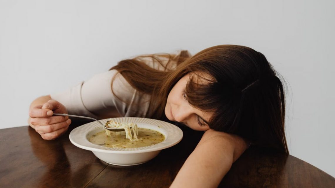 A woman resting her head on the table while holding a spoon and eating soup, conveying fatigue.
