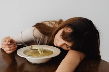 A woman resting her head on the table while holding a spoon and eating soup, conveying fatigue.