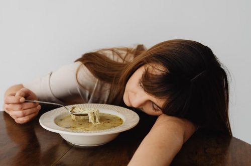 A woman resting her head on the table while holding a spoon and eating soup, conveying fatigue.