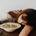 A woman resting her head on the table while holding a spoon and eating soup, conveying fatigue.